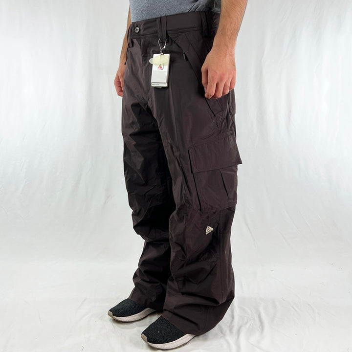 2006 Deadstock Vintage Nike ACG Cargo Ski Pants in Brown with Nike ACG branding. Loose fit, Cargo trousers look with plenty of pockets including zip pockets to inner thighs. Storm fit technology. Fleeced inner layer and boot gaiters. Belt loops for belt adjustment. - Materials: Nylon/Polyester - Colour: Brown Brand New with Tags - Size on Tag: Medium Measurements: Length: 42 Inches Inseam: 32 Inches Waist: GB 31/33 - Size on Tag: Large Measurements: Length: 43 Inches Inseam: 32.5 Inches Waist: GB 34/36