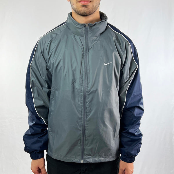 Y2K Deadstock Vintage Nike Swoosh Windbreaker Jacket in Grey. Nike Swoosh logo to chest. Pockets to front. Zip closure to jacket. Adjustable cord to waist. Hood hidden in hood compartment behind neck. Adjustable cord to hood. - Materials: Nylon/Polyester - Colour: Grey