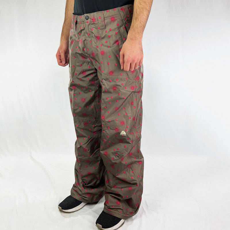 Y2K Women's Deadstock Vintage Nike ACG Cargo Ski Pants in brown with Nike ACG branding. Pink design all over. Storm-Fit technology with fleeced inner layer. Zip pockets to sides. Boot gaiters. Belt loops for belt adjustment. - Materials: Polyester - Colour: Brown Brand New with Tags
