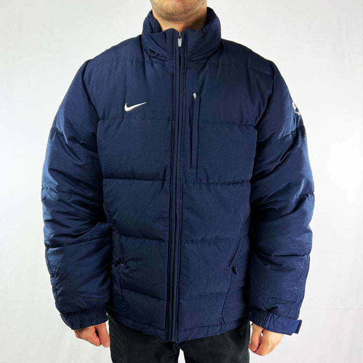 Y2K Deadstock Vintage Nike puffer jacket in navy blue. Embroidered Nike swoosh to chest and Nike Football logo to arm. Zip pockets. Zip closure to jacket. Adjustable cord to waist. Hidden hood in hood compartment behind neck. Adjustable cords to hood. Zip under arms. - Materials: Body: 100% Nylon Fill: White Duck Down Feathers & Down