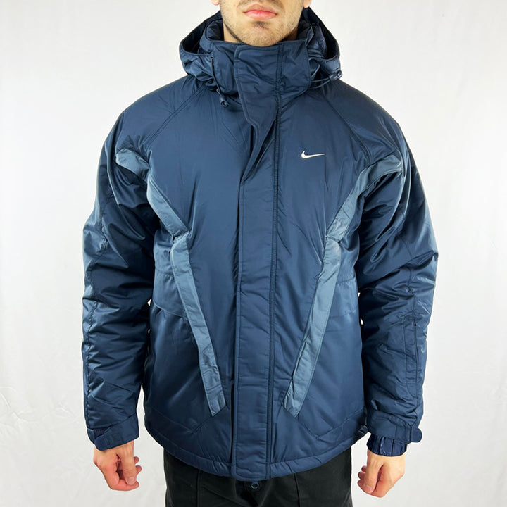 Y2K Deadstock Vintage Nike swoosh puffer jacket in navy blue. Embroidered Nike swoosh to chest. Zip pockets to front. Zip closure to jacket. Inner pocket. Removeable hood. Adjustable cord to hood. Padded. - Materials: Polyester - Colour: Navy Blue Brand New with Tags - Size on Tag: Small Measurements: Pit to Pit: 22.5 Inches Length: 27.5 Inches